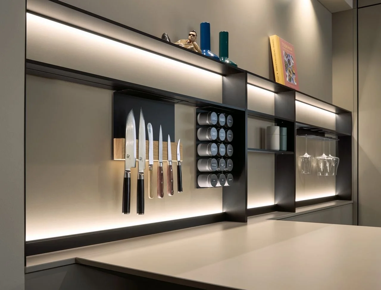 Modern kitchen wall with knives, spice containers, wine glasses, and decorative vases, illuminated by built-in lighting.