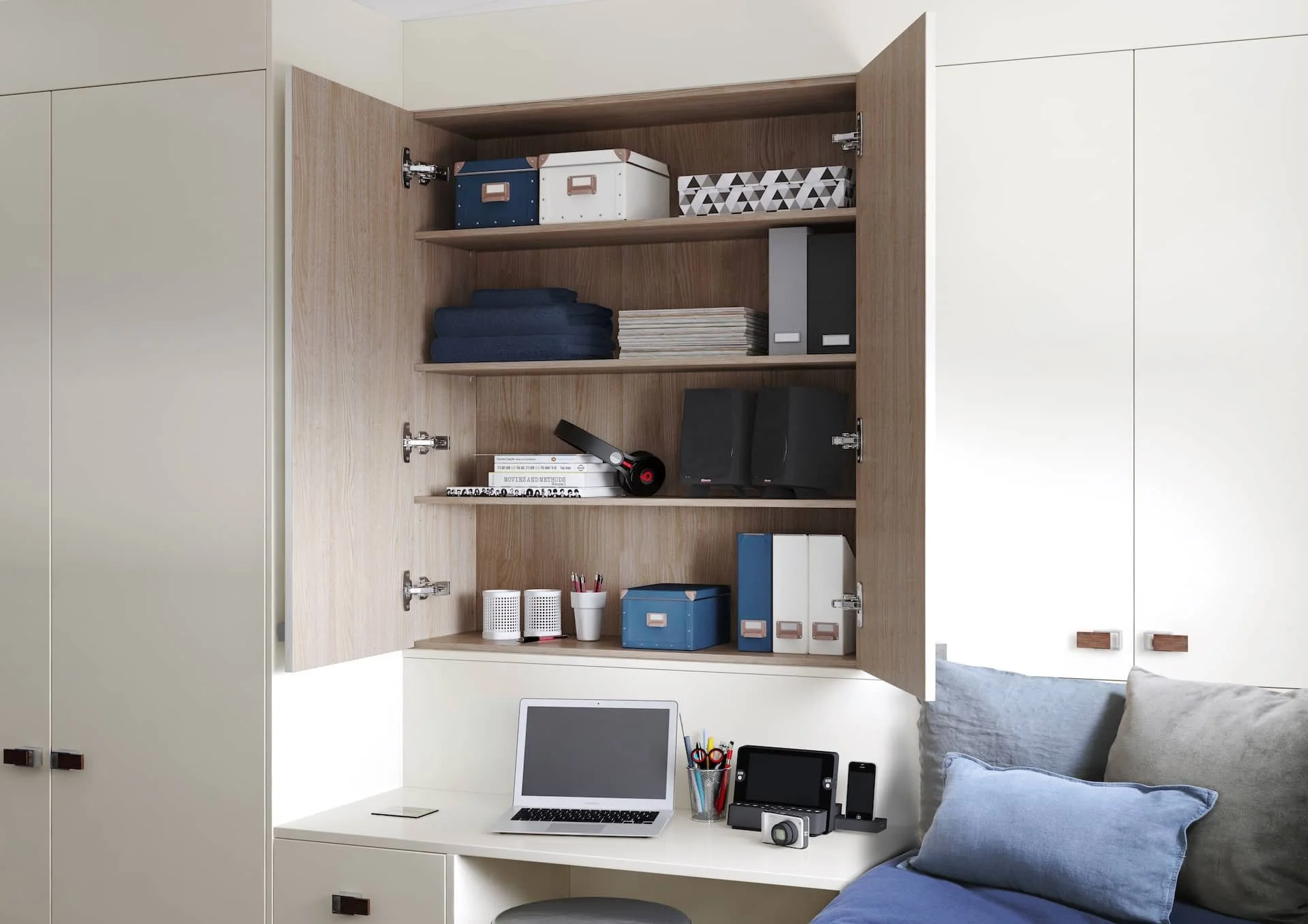 Open wooden cabinet with shelves above a white desk, containing boxes, folders, books, and a headset, with a laptop, camera, and stationery on the desk, and pillows on a bed beside it.