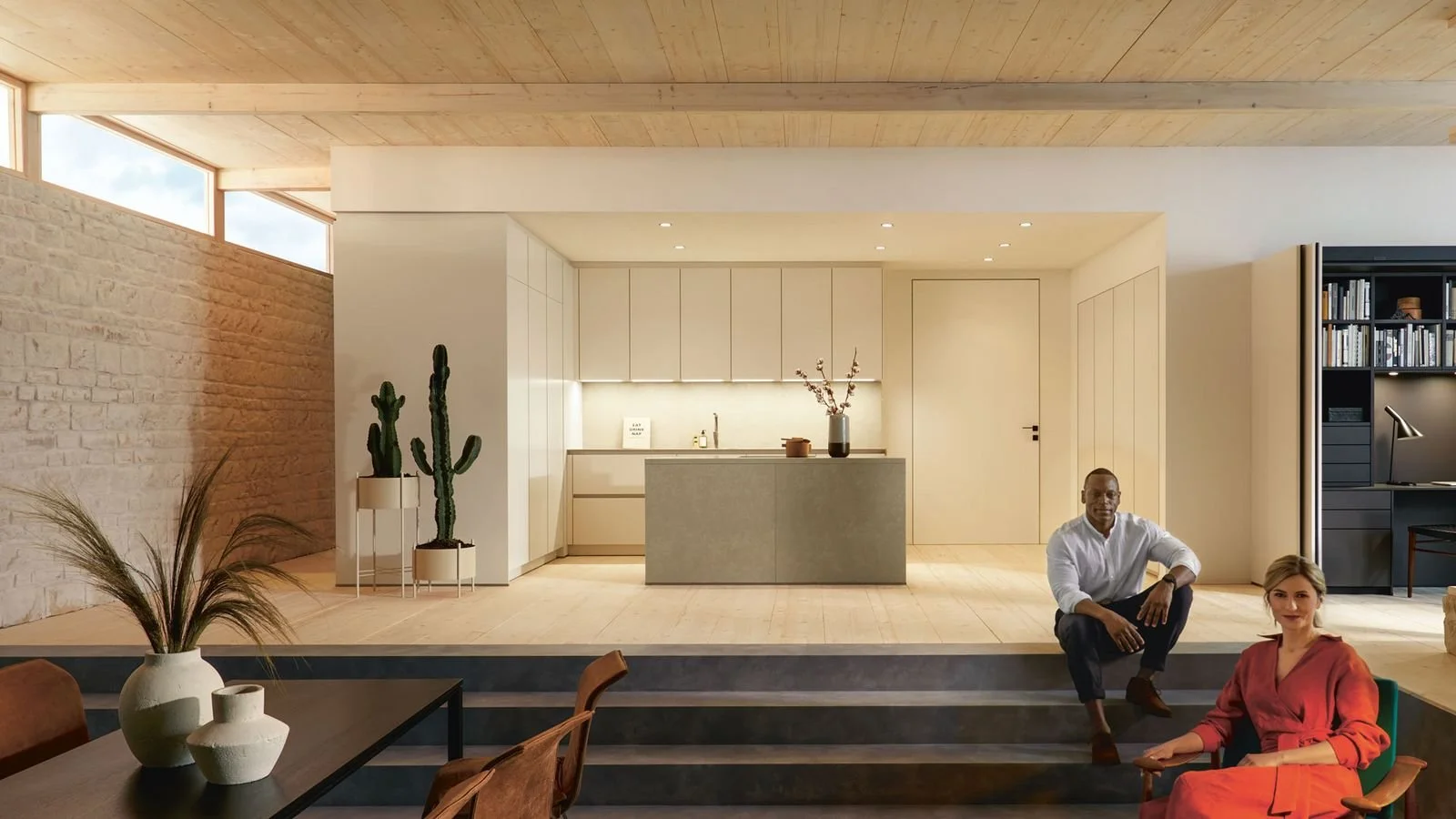 Modern open-concept living space with a kitchen area, two people sitting on stairs, and decorative plants.