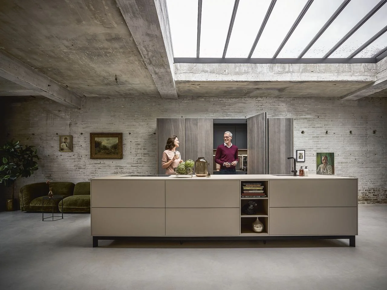 Two people are standing and talking in a modern kitchen with exposed brick walls, a skylight ceiling, and a large island with decorative objects.