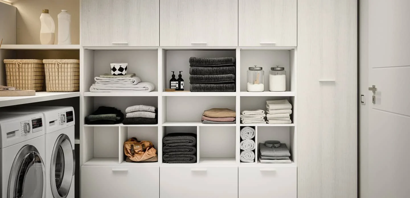 Laundry room with shelves containing folded towels, baskets, bottles, and laundry supplies, with washing machines on the left.