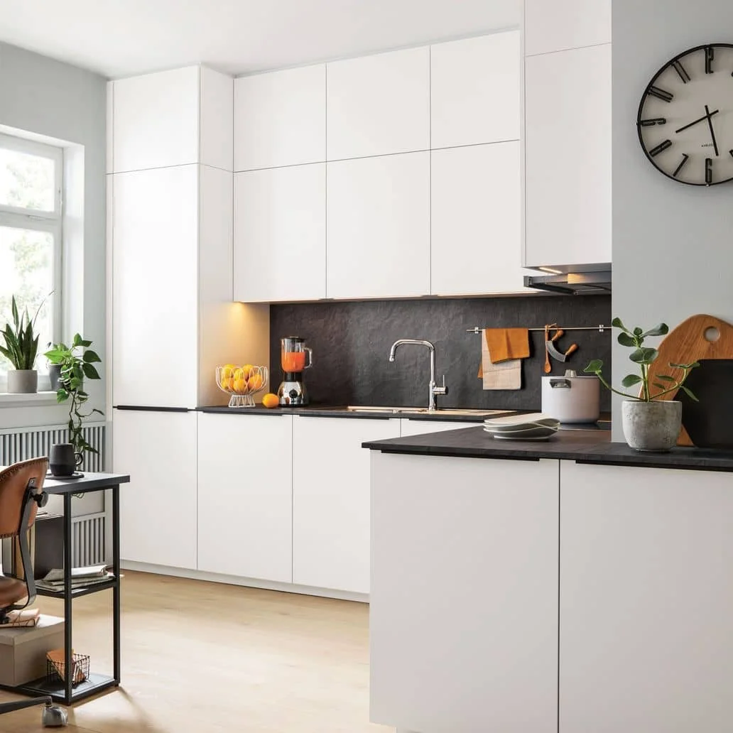 Modern kitchen with white cabinets, black countertop, and black backsplash. A window with plants, a clock on the wall, and various kitchen items including a blender, bowl of fruit, and potted plants