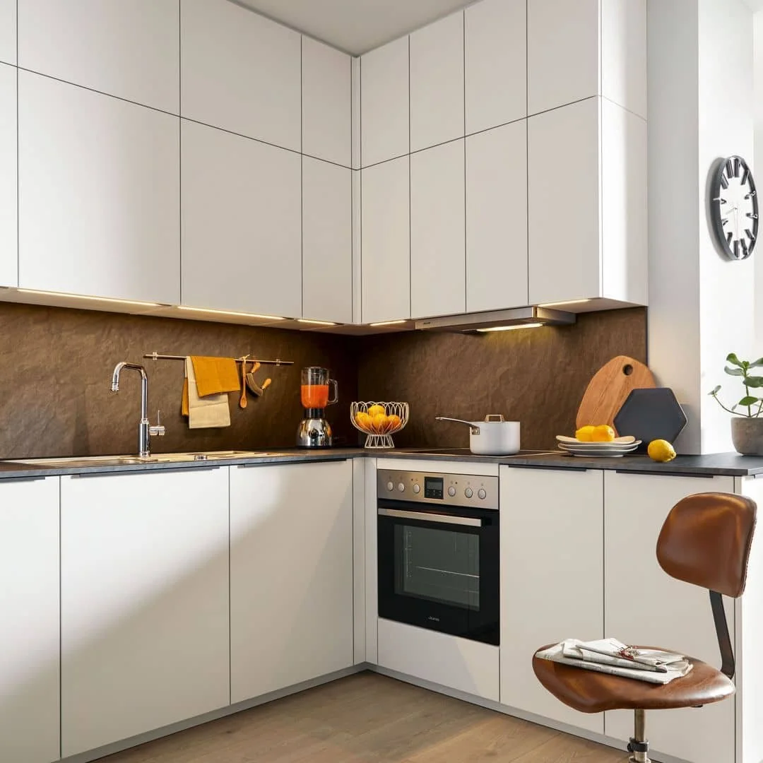 Modern kitchen with white cabinets, dark countertop, brown backsplash, blender with orange drink, fruit bowl, white pot, cutting board, and a plant on the window sill.