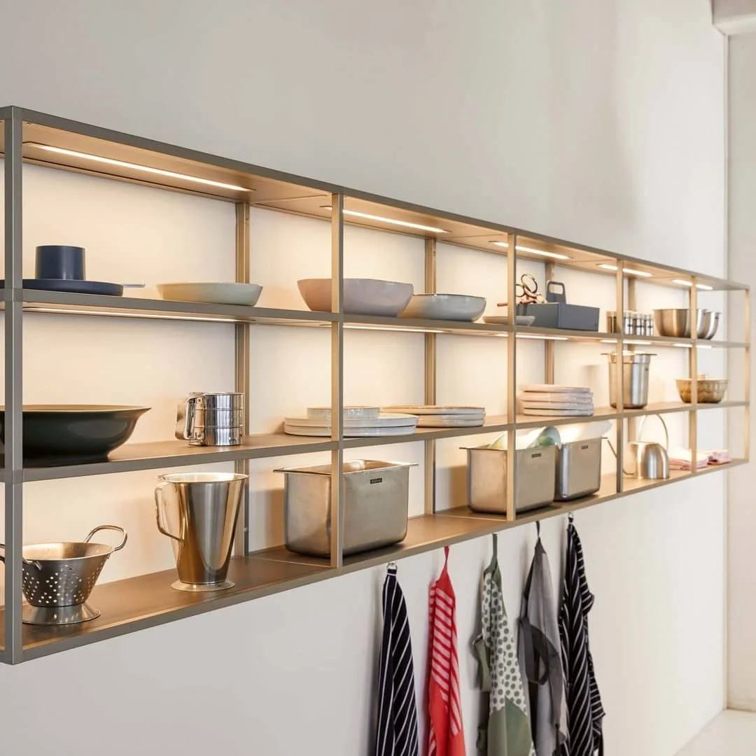 Wall-mounted open shelving unit with kitchenware including bowls, plates, utensils, and towels. Items displayed are primarily in neutral colours with some metallic accents.