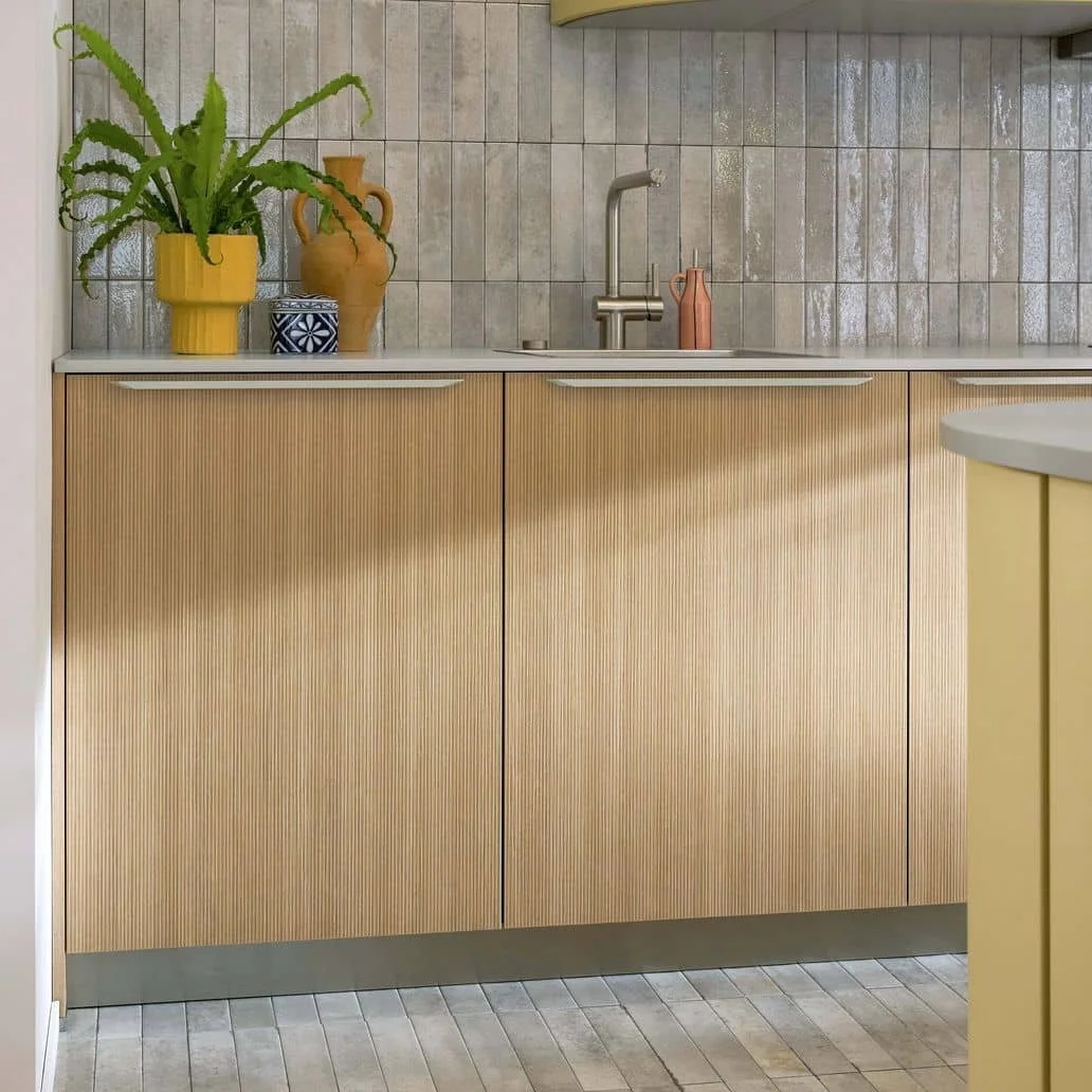 Kitchen counter with yellow cabinets, a plant in a yellow pot, a blue and white patterned jar, and a large brown vase; tiled backsplash and a stainless steel faucet.