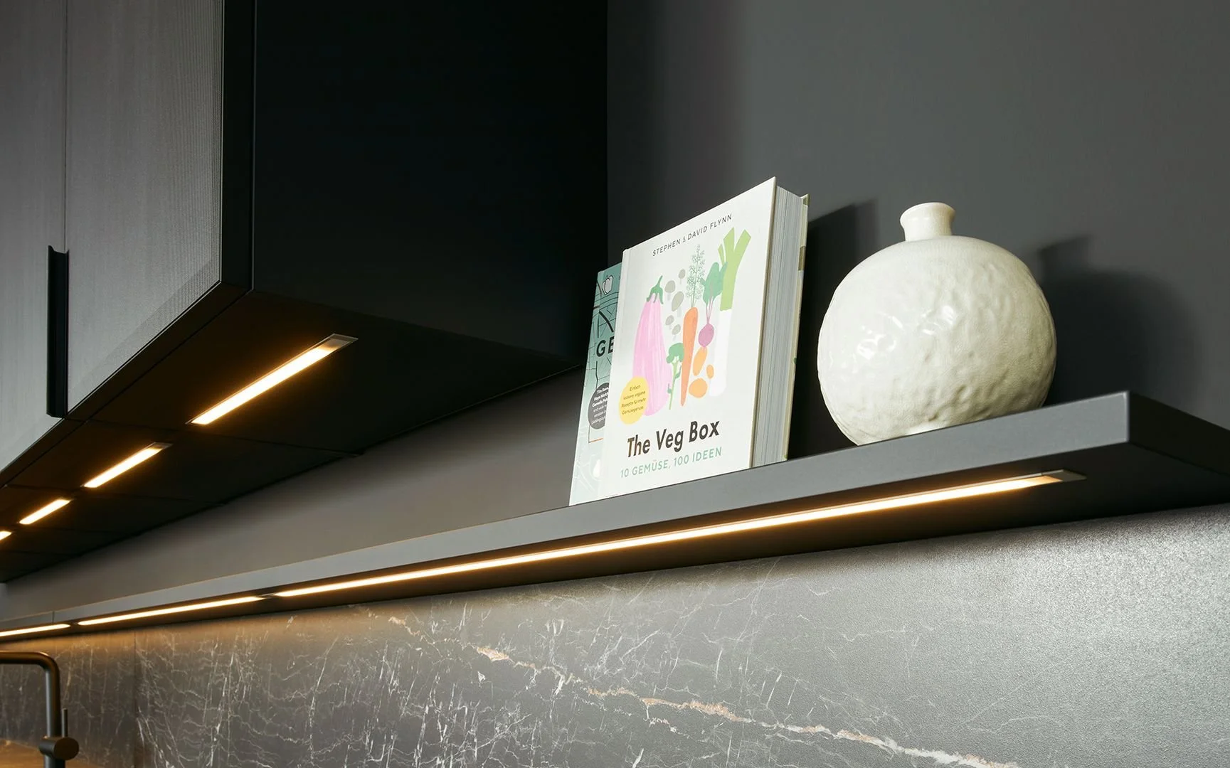 A modern kitchen shelf holding a cookbook titled "The Veg Box" and a textured white ceramic vase.