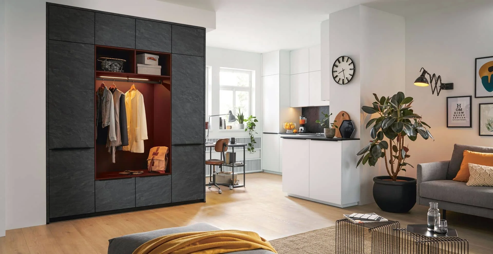 Modern living room with an open closet featuring hanging clothes, a small kitchen area with plants on the window sill, a large indoor plant, wall art, and a gray sofa with pillows.