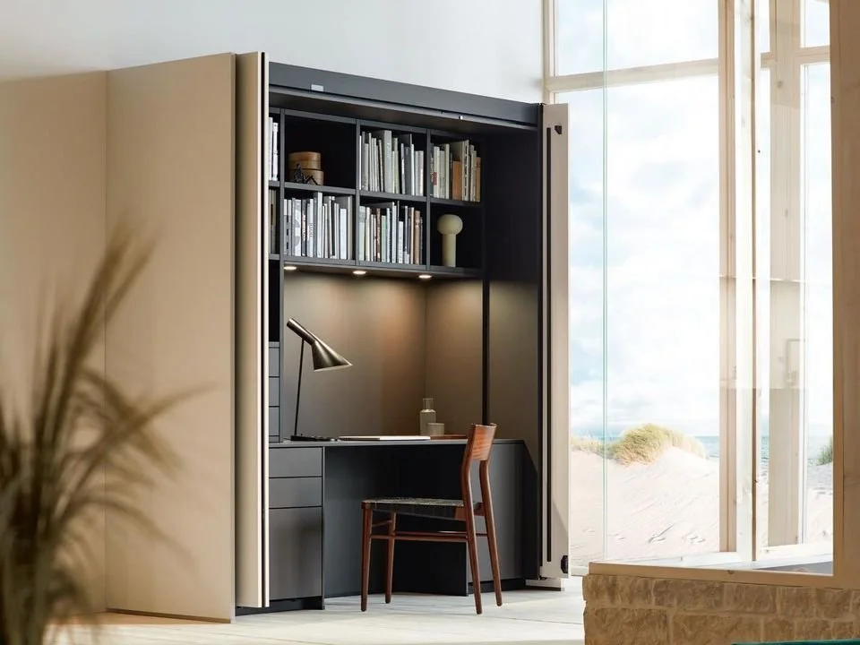 Modern built-in home workspace with a desk, chair, and bookshelf inside a hidden cabinet, near large windows overlooking sandy dunes.