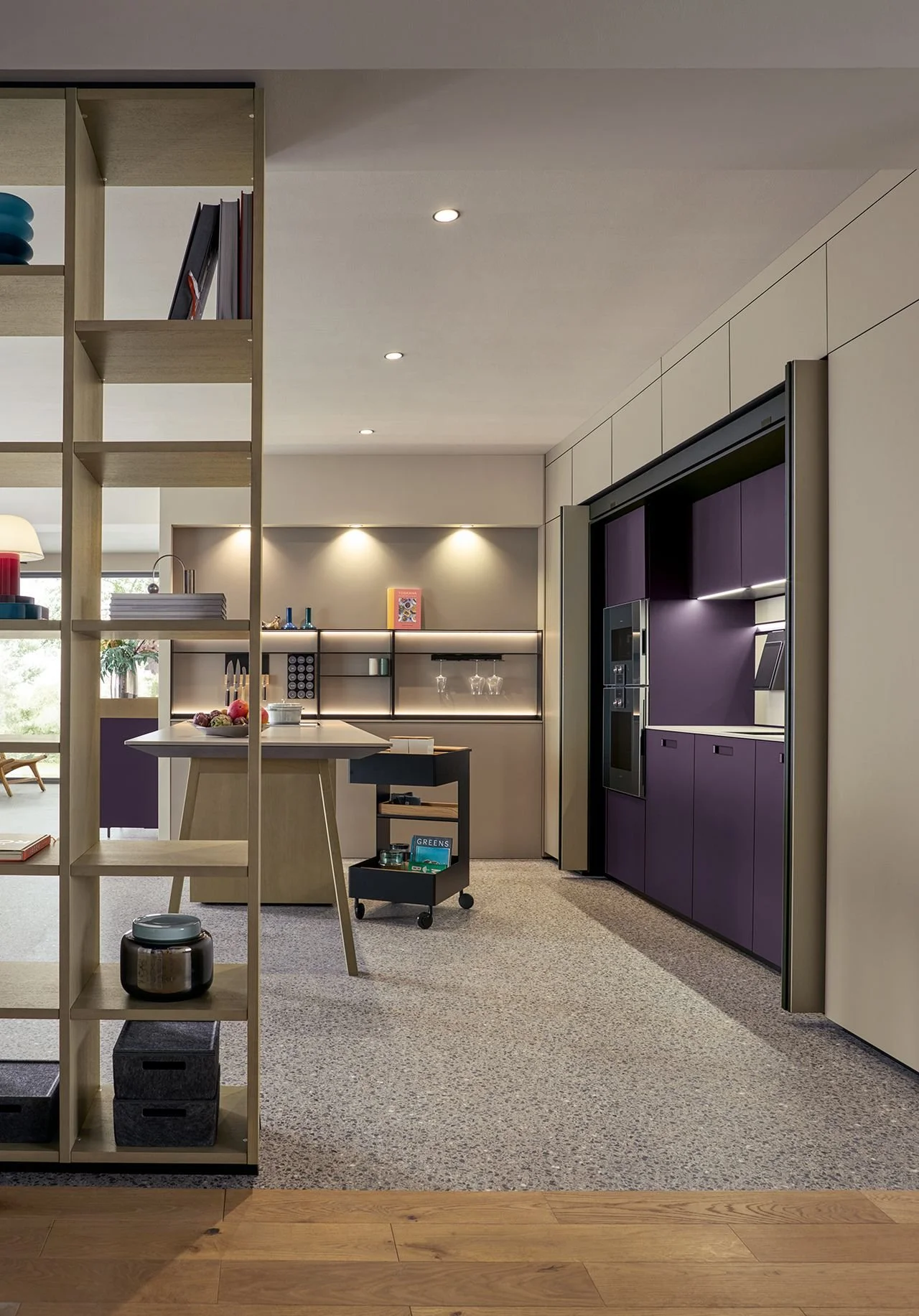 Modern kitchen with purple cabinets, beige walls, and minimalist decor, featuring a kitchen island with fruit, a rolling cart with books, and a open shelving unit with glasses and decorative items.