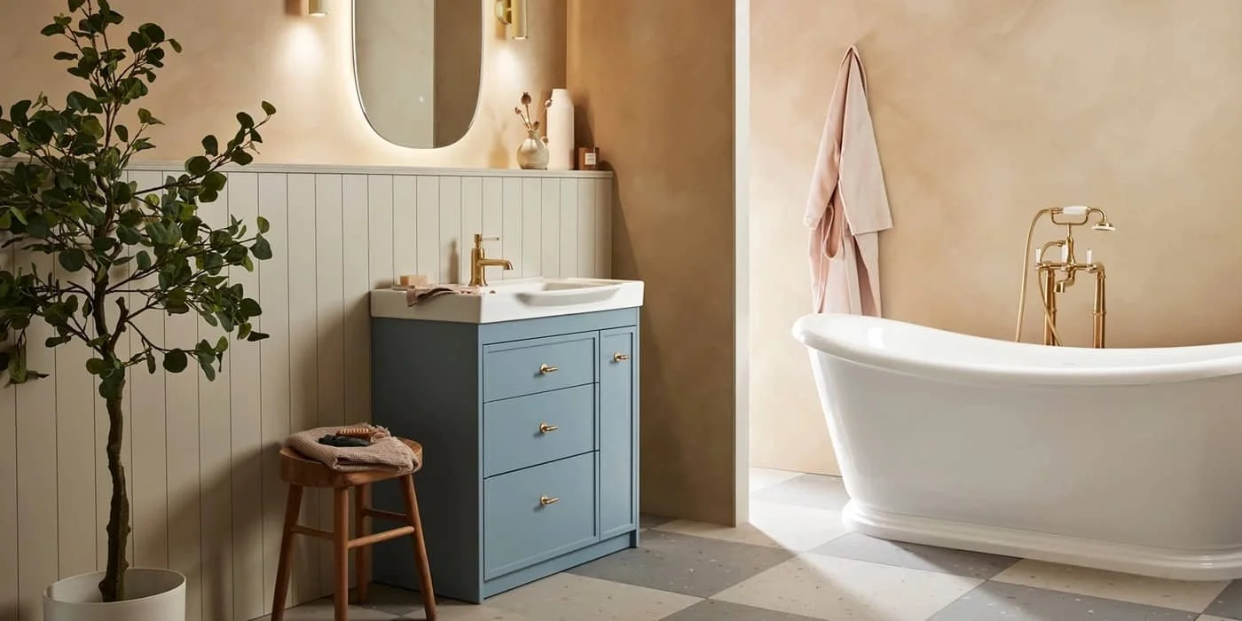 A traditional bathroom with a freestanding bathtub, a blue vanity with gold hardware, a round mirror with backlit, a small wooden stool with towels, and a potted plant.