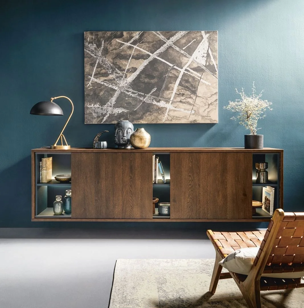 A modern living room with a teal wall, a wooden Schüller sideboard with open shelves, a black and gold desk lamp, decorative objects, a large abstract wall art, a potted plant, and a wooden chair with a cushion.