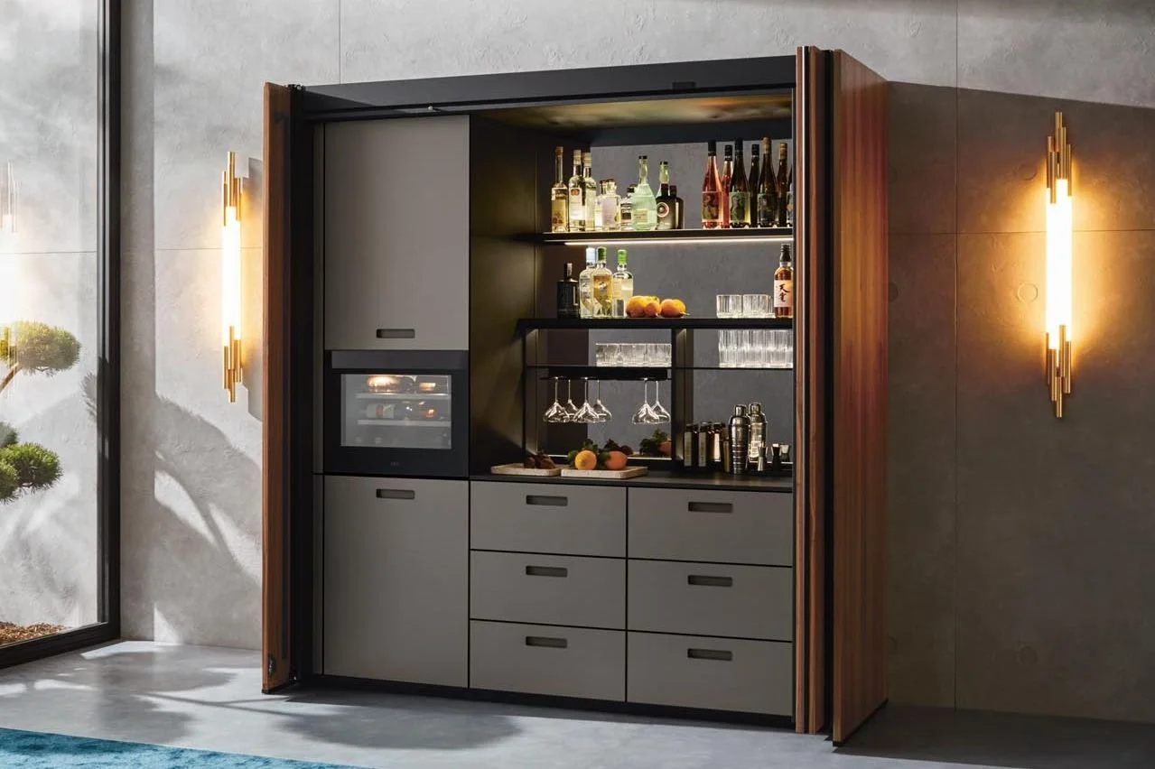 A modern built-in drinks bar inside a cabinet with open sliding doors, containing bottles of alcohol, glasses, and bar accessories.