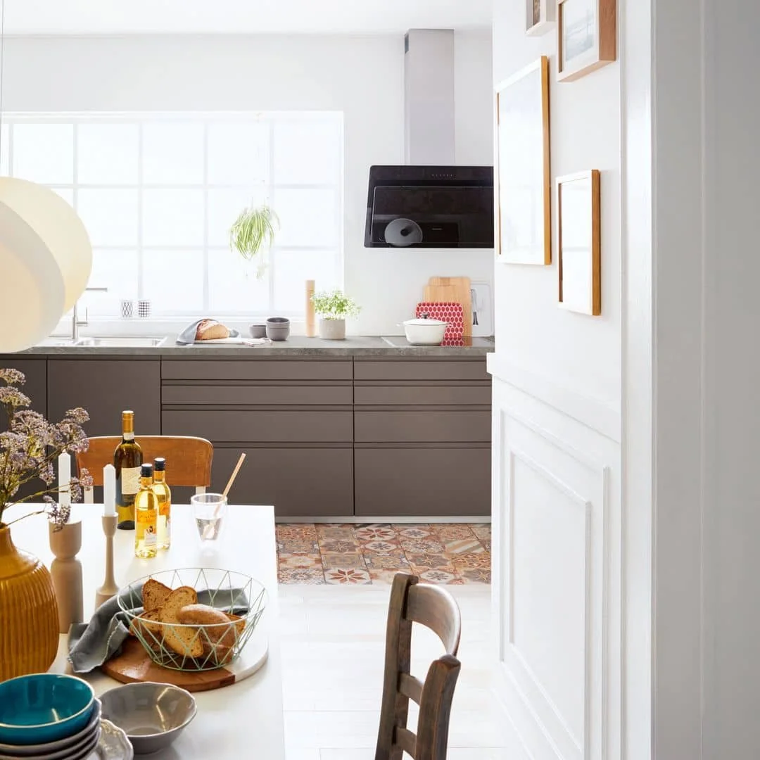 Bright, modern kitchen with gray cabinets, a white countertop, a large window, and a dining table with bread, flowers, and drinks.