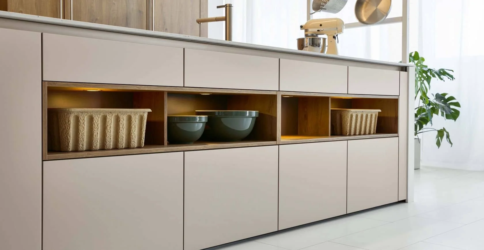 Modern kitchen cabinet with open shelving, displaying ceramic bowls and woven baskets near a plant, in a bright light-filled space.