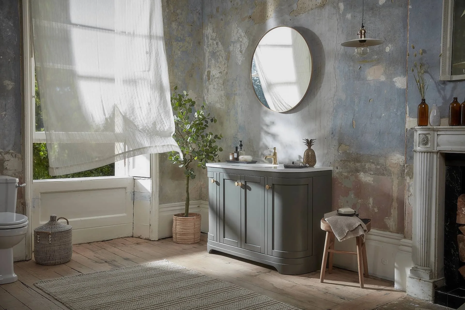 Traditional bathroom with distressed wall paint, a round mirror, a grey vanity, a potted plant, a woven basket, a window with sheer white curtain, and a small stool with towels.