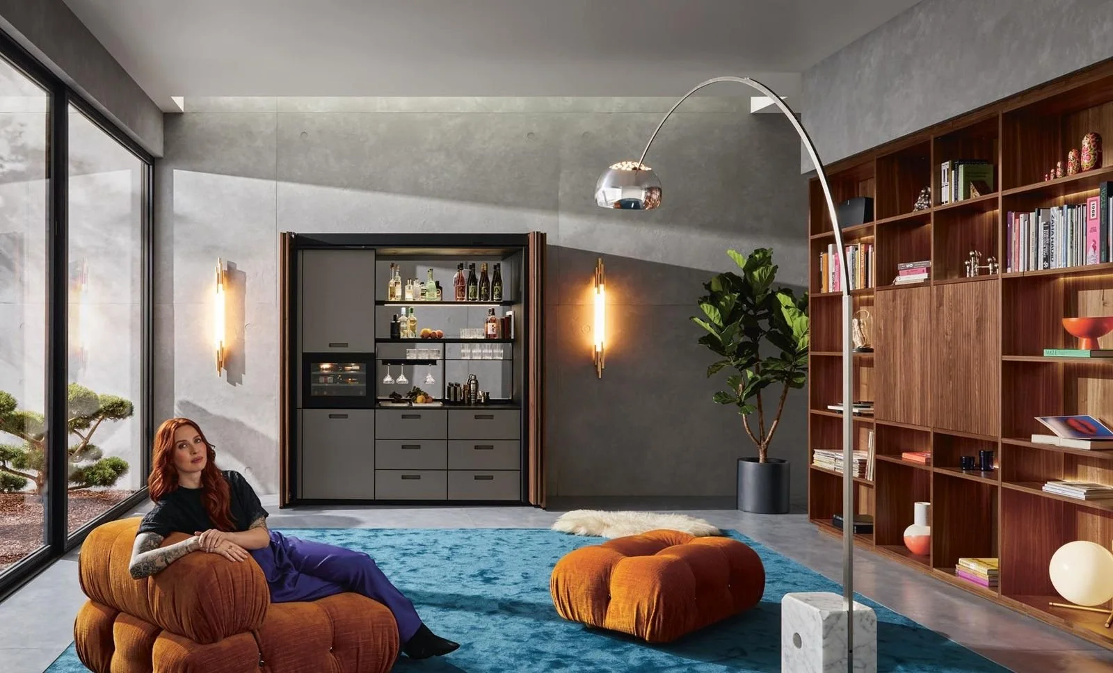 Interior view of a modern living room with a woman sitting on an orange sofa, a large blue rug, a wooden bookshelf filled with books and decorative objects. Large sliding glass doors reveal an outdoor garden.