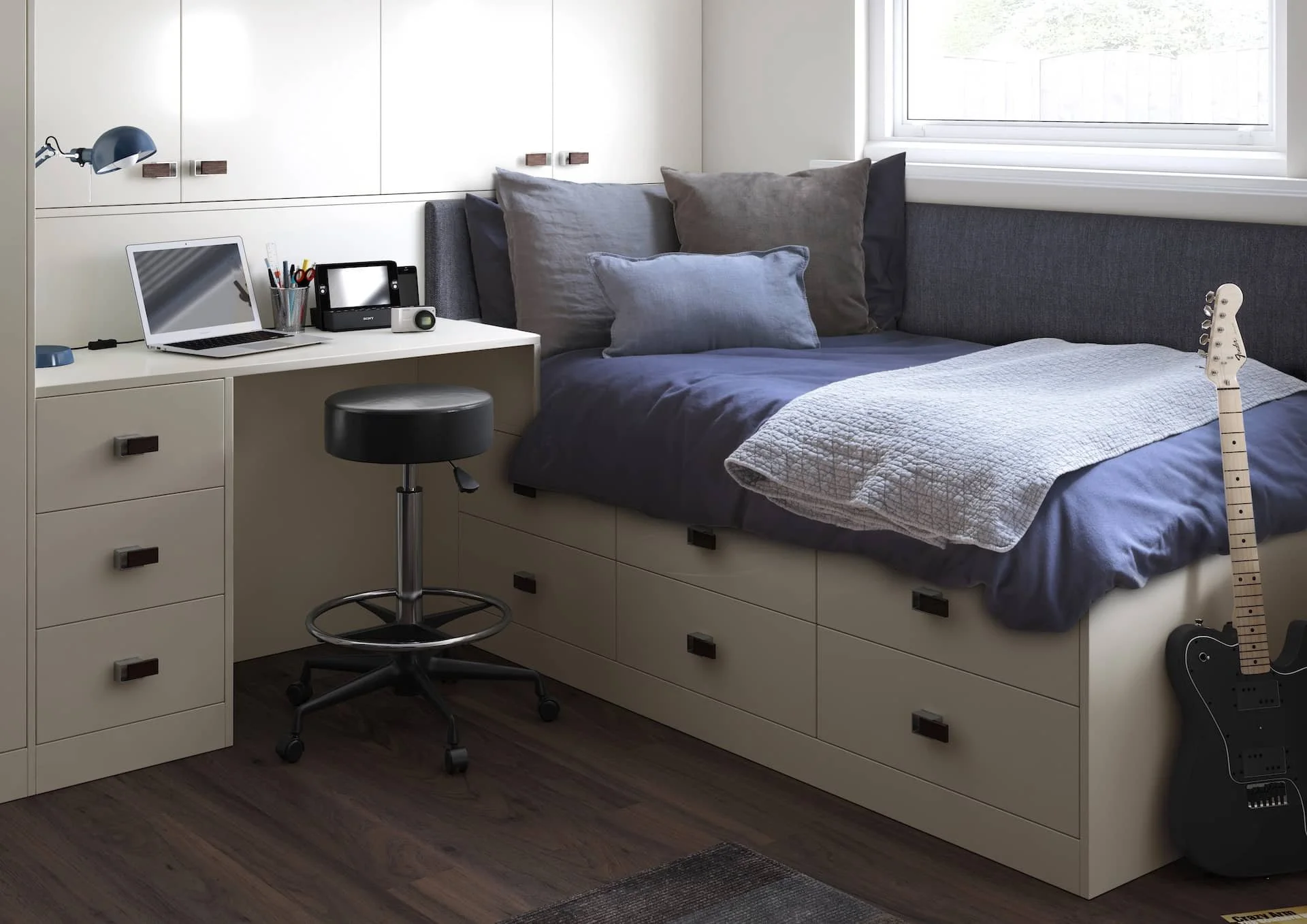 A small bedroom with a bed built into a white storage unit, a black electric guitar leaning against the bed, and a white desk with a laptop, printer, and stationery.