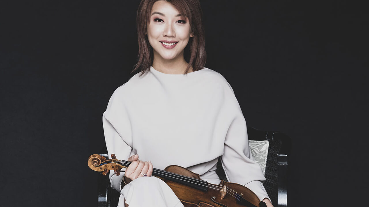 Mujer sonriendo, con cabello castaño, vestida con blusa blanca, sosteniendo un violín en sus manos, con fondo negro