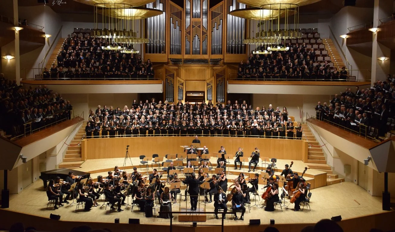 Un coro grande en el fondo, una orquesta en el escenario y un director de orquesta en el centro. La sala es de madera y tiene iluminación cálida.