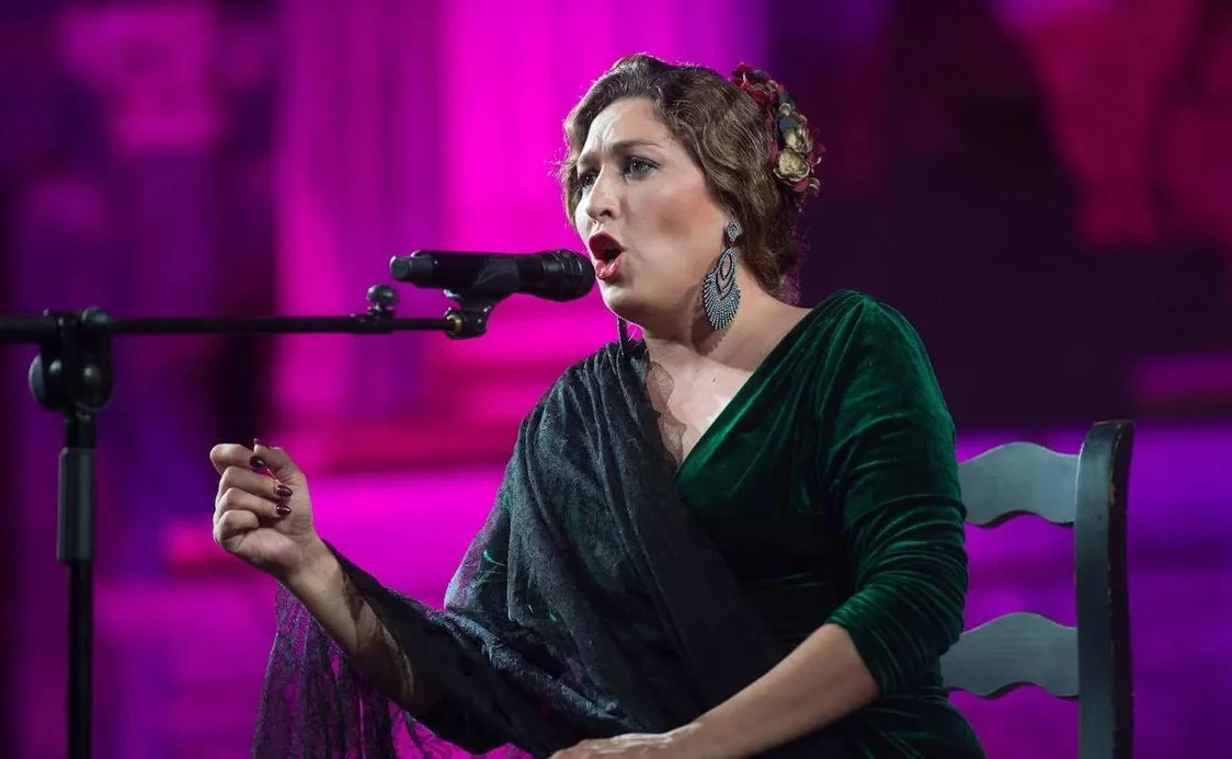 Mujer cantando frente a un micrófono, vestida con vestido verde y con accesorios en el cabello, fondo rosa brillante.