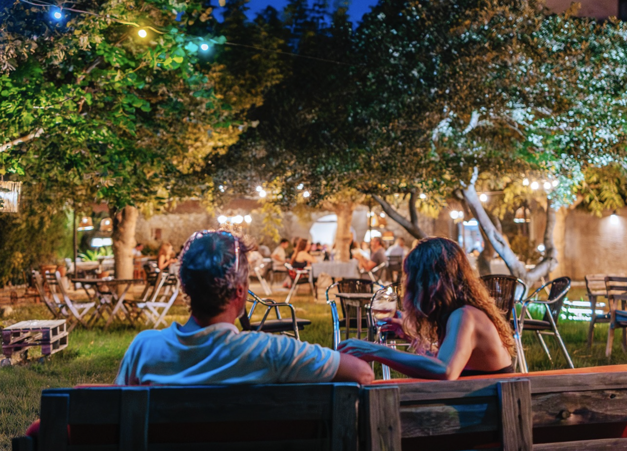 Pareja sentada en un área al aire libre con árboles y luces decorativas en la noche, en un lugar que parece ser un restaurante o café con más personas en el fondo.