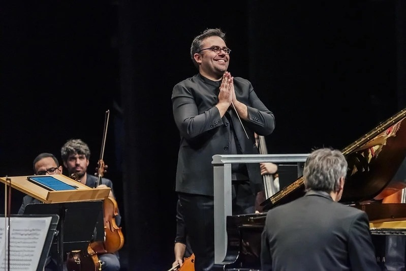 Un director de orquesta sonriente con las manos juntas en señal de agradecimiento, rodeado por músicos con instrumentos en un escenario.