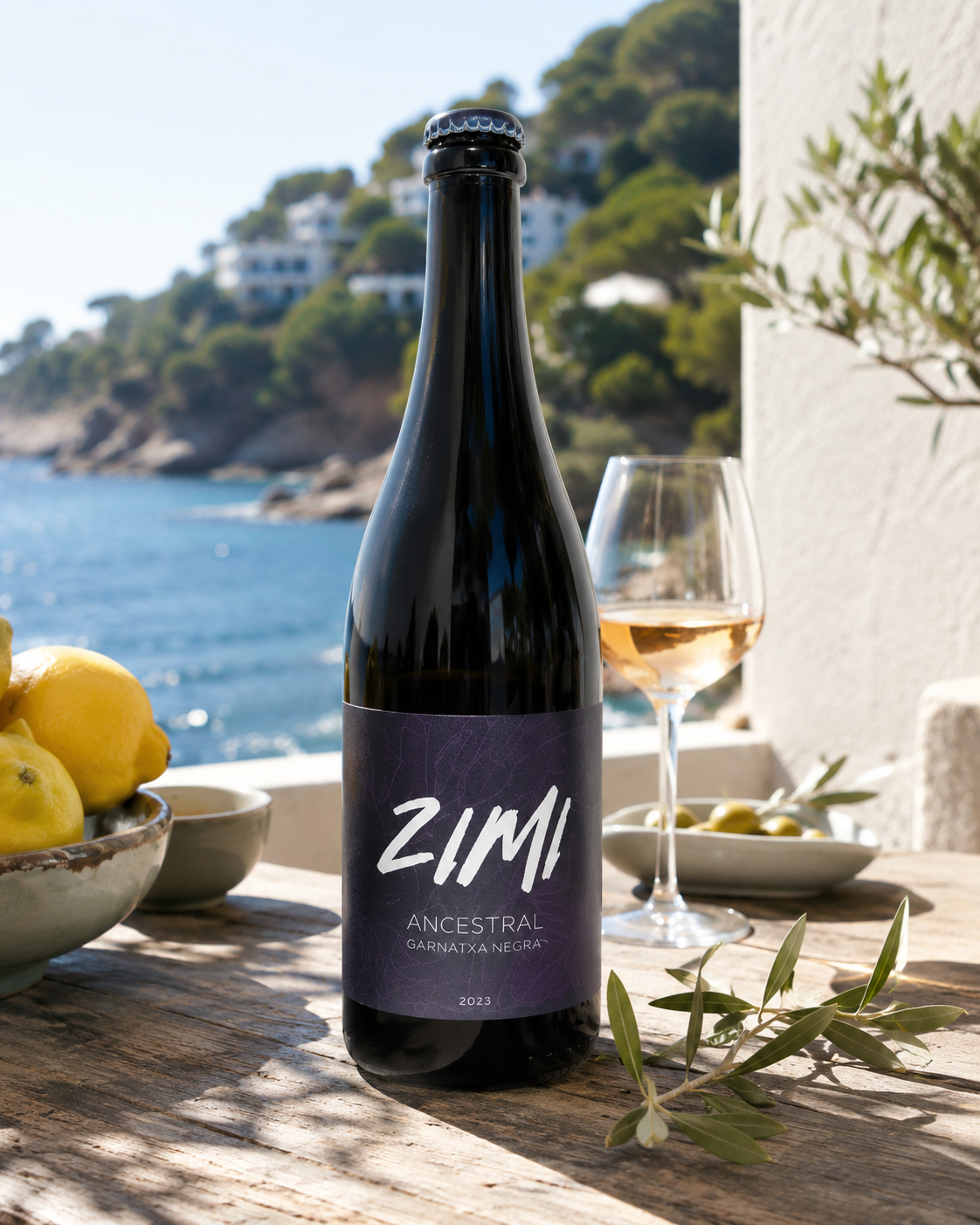 Botella de vino Zimi Ancestro Garnatxa Negra 2023 en una mesa de madera con vista al mar, acompañada de un vaso con vino rosado, limones en un tazón, aceitunas y ramas de olivo.