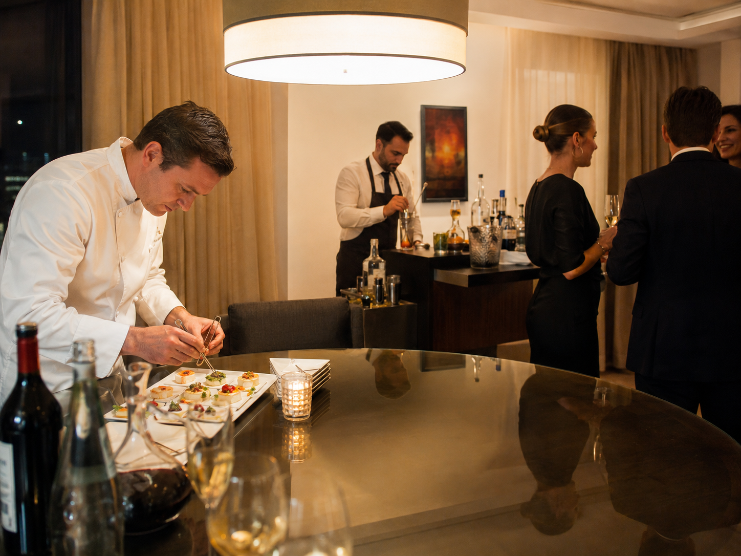 Chef decorando canapés en una mesa de comedor elegante con varios invitados sociales en el fondo en una habitación con iluminación cálida.