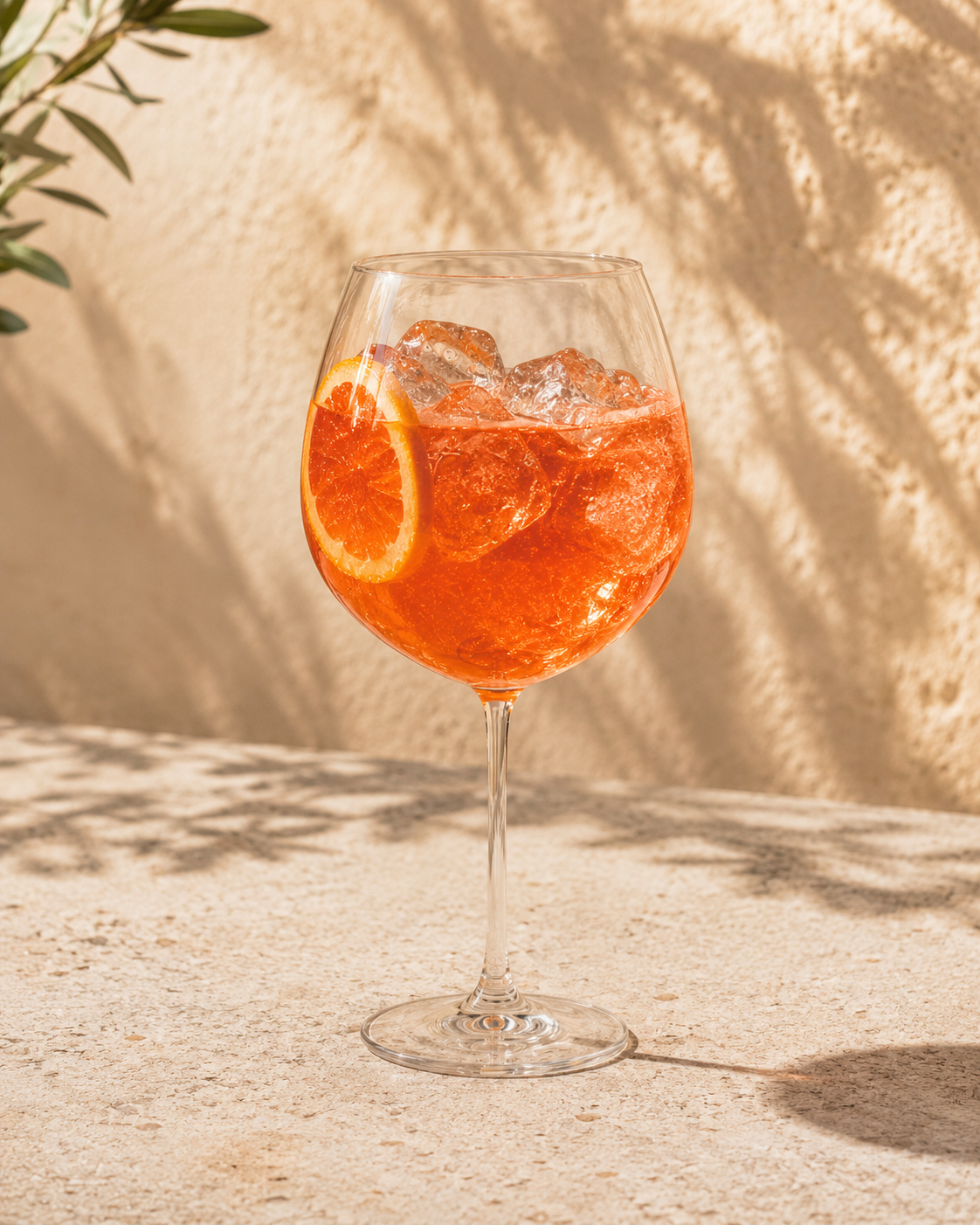 Copa de vidrio con bebida de color naranja, hielo y una rodaja de naranja decorativa, sobre una superficie de piedra con sombra y fondo de pared beige con sombra de hojas.