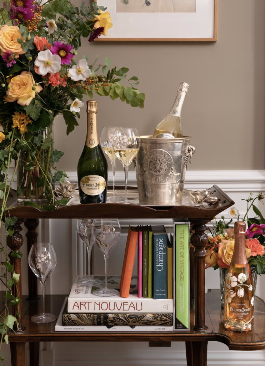 Flores variadas en un florero, botellas de champagne, copas y libros en una mesa de madera.