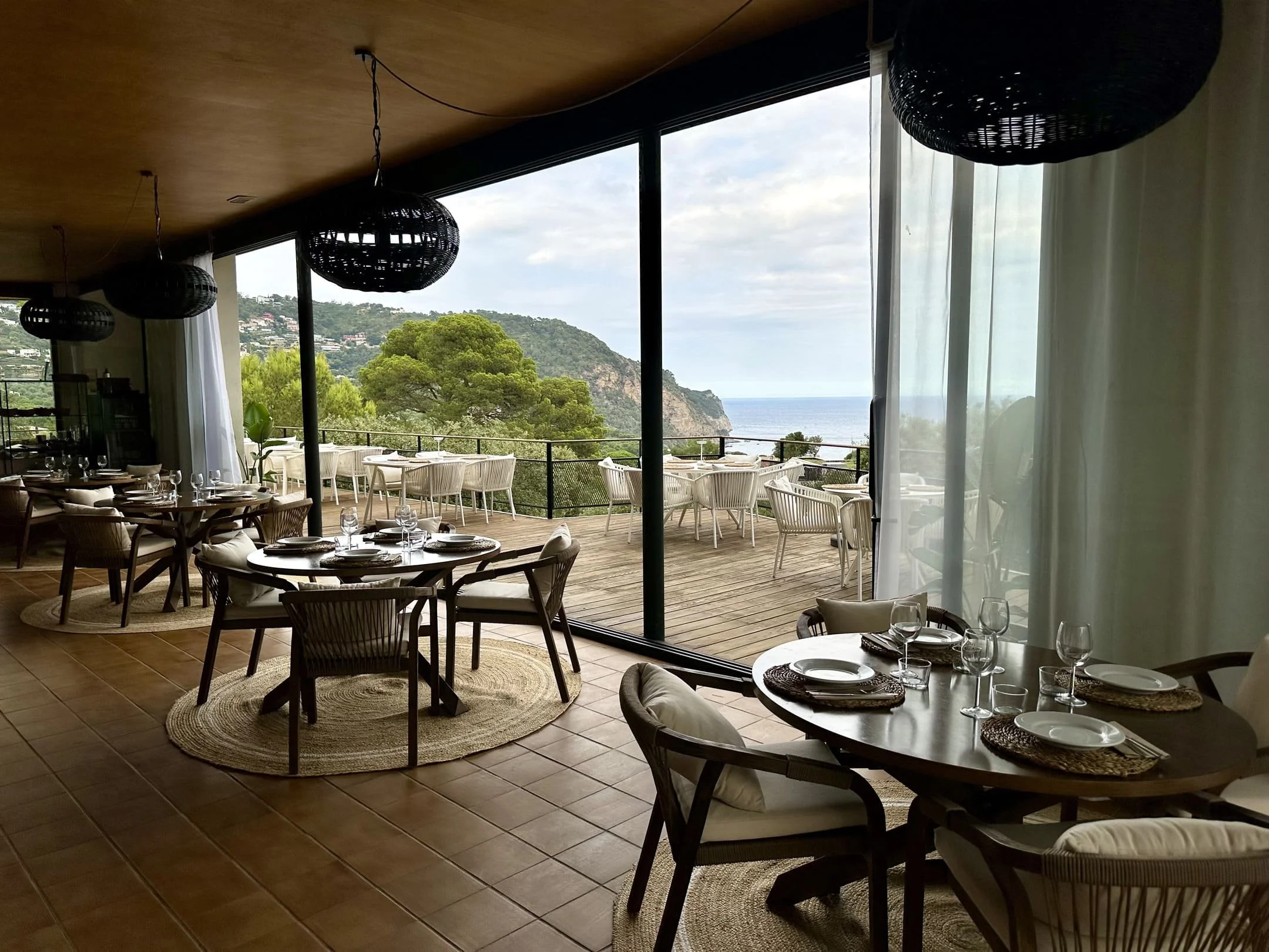 Vista desde el interior de un restaurante con mesas y sillas, al fondo ventanas grandes que muestran una vista de un área con árboles, una colina y el mar, días nublados.