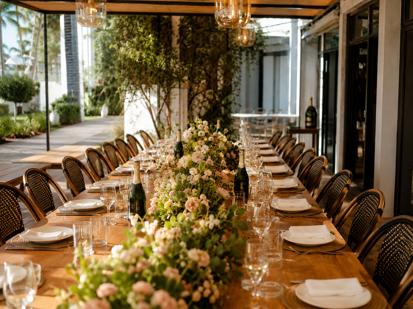 Mesa larga decorada con flores y botellas de champagne en un espacio abierto con luz natural y plantas verdes.