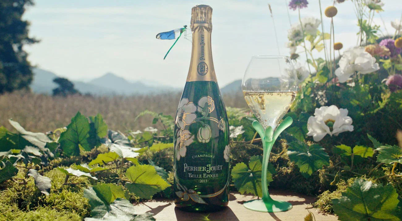 Botella de champagne Perrier-Jouët decorada con flores y una copa con vino espumoso, en un entorno natural con flores y un paisaje de montañas al fondo.