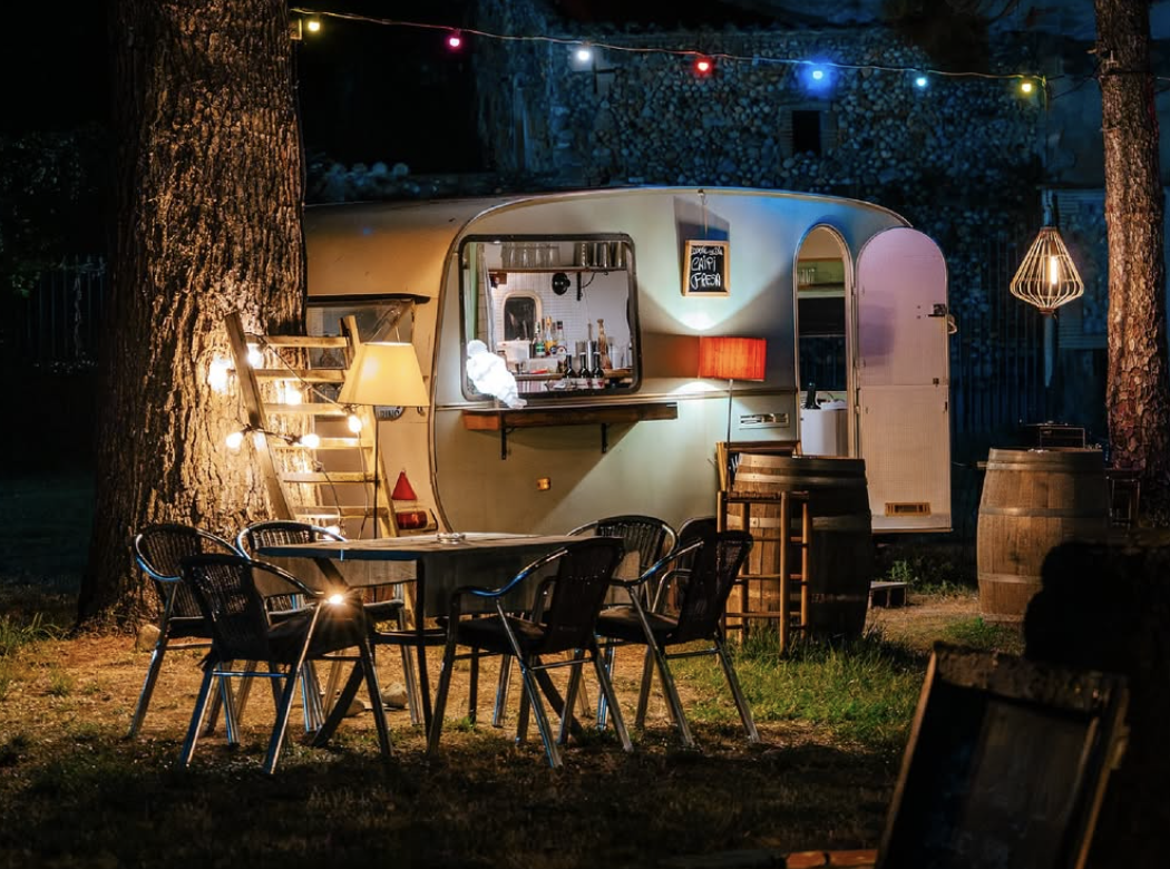 Cabaña de estilo vintage en un espacio al aire libre, decorada con luces de colores y lámparas, con un área de bar y mesas y sillas de patio.