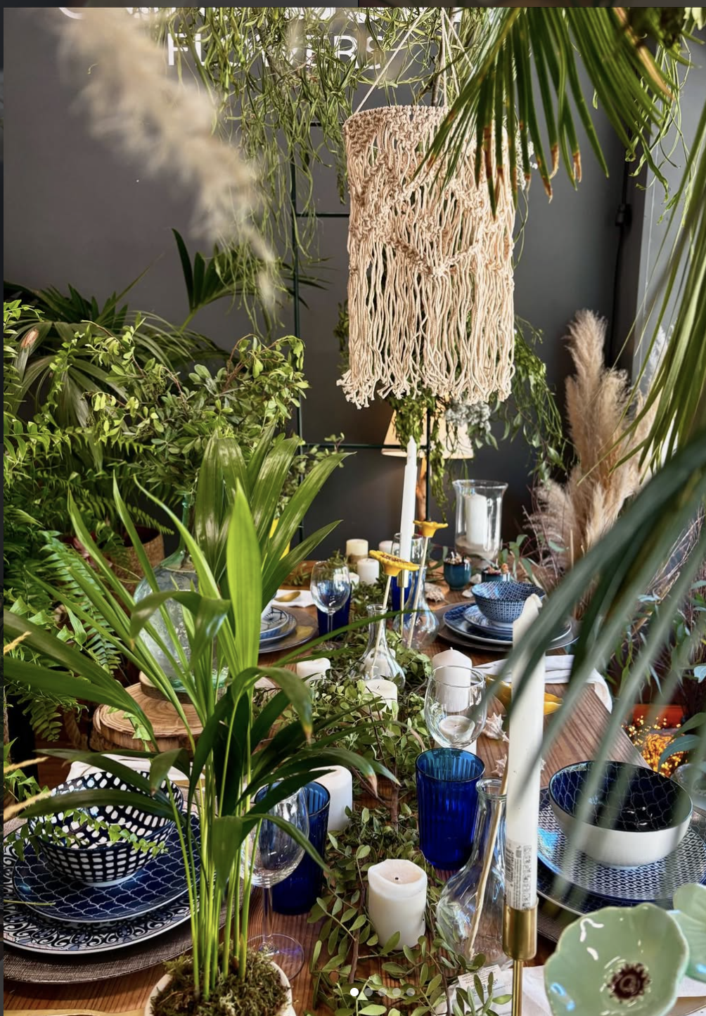 Mesa decorada con platos, vasos y velas, rodeada de muchas plantas verdes y plantas con flores, en un ambiente elegante y natural.