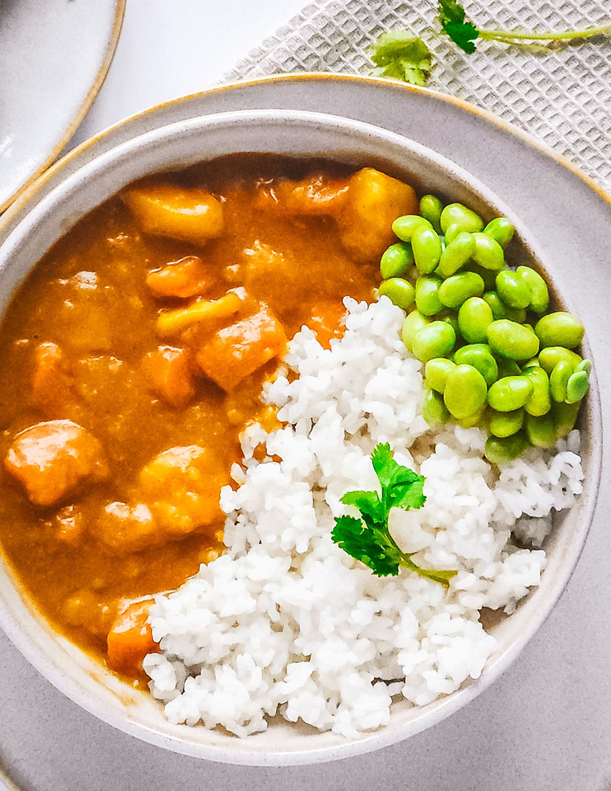 Beige bowl of vegetarian Japanese curry with rice and edamame beans, garnished with a single coriander leaf, set on a beige plate with a napkin on a white tiled background.