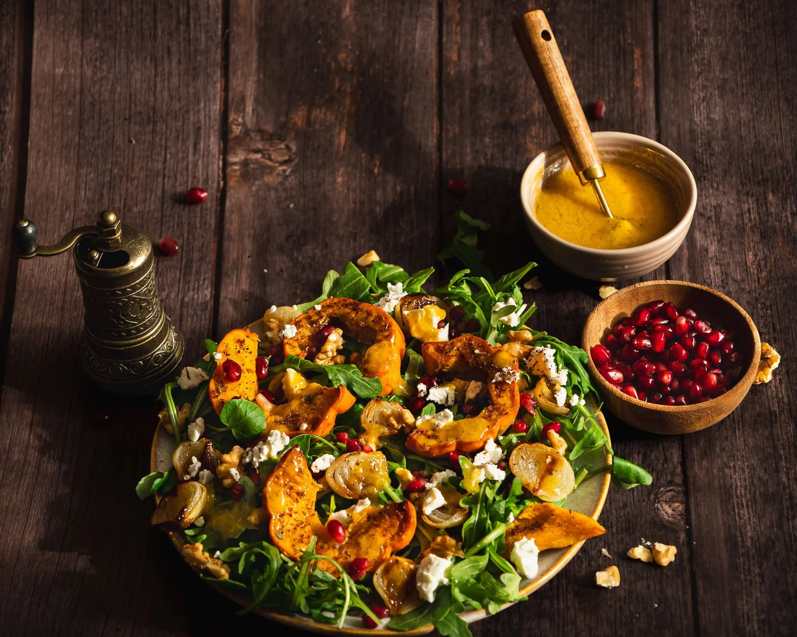 Side-angle view of roasted munchkin pumpkin and baby onion salad on a cream ceramic plate, layered with rocket, crumbled feta, walnuts and pomegranate, with dressing and a brass pepper mill in the background.