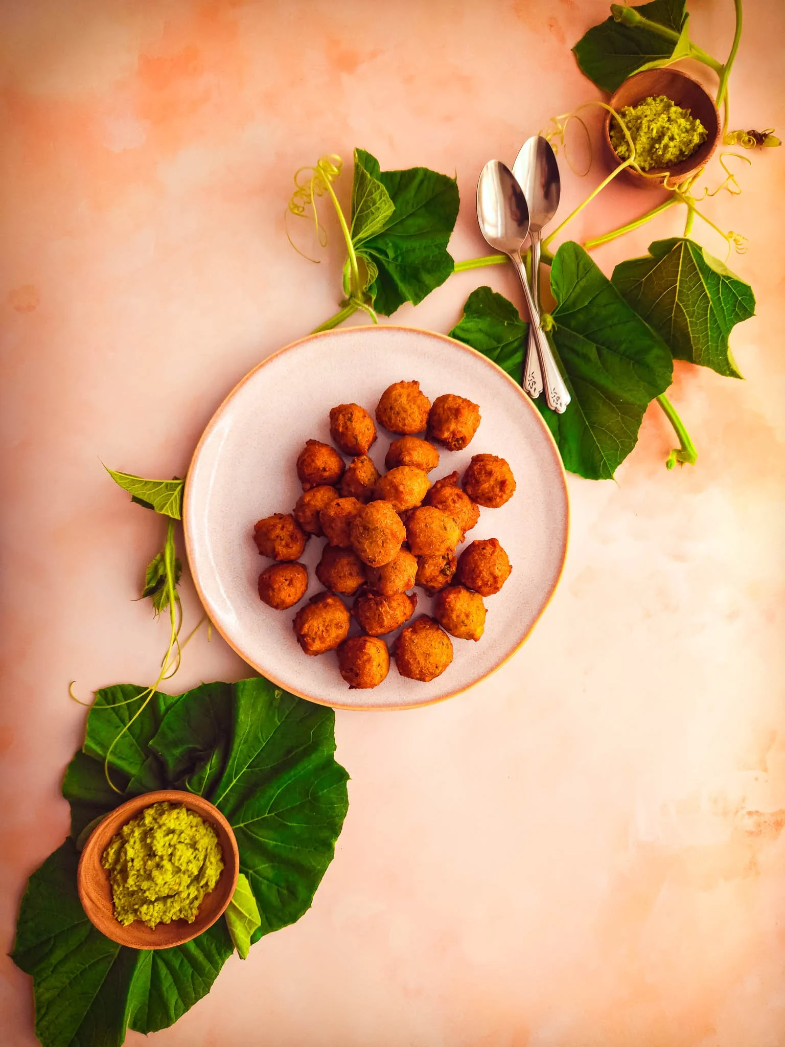 Diagonal arrangement of dudhi pakoda on a white plate, fresh dudhi leaves laid diagonally across the plate, green chutney bowls bottom-left and top-right, with two spoons placed alongside