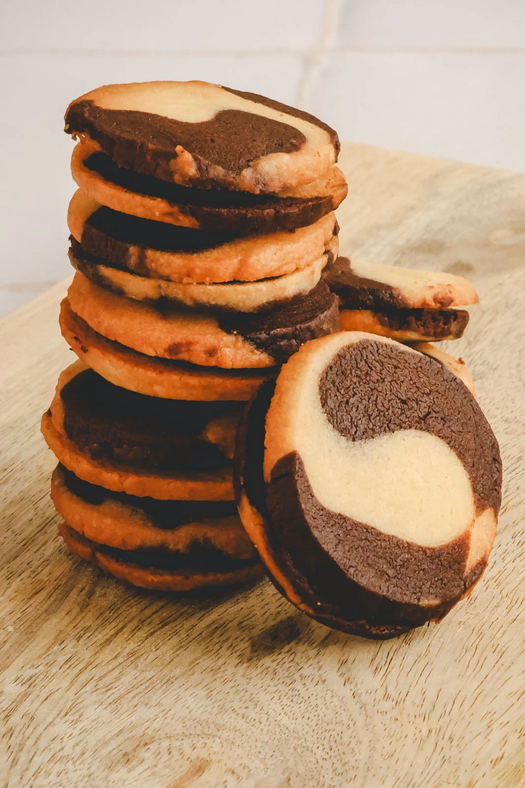 Marble sablé sandwich cookies filled with dark chocolate ganache, arranged on a wooden board with one cookie propped beside the stack in soft natural light.