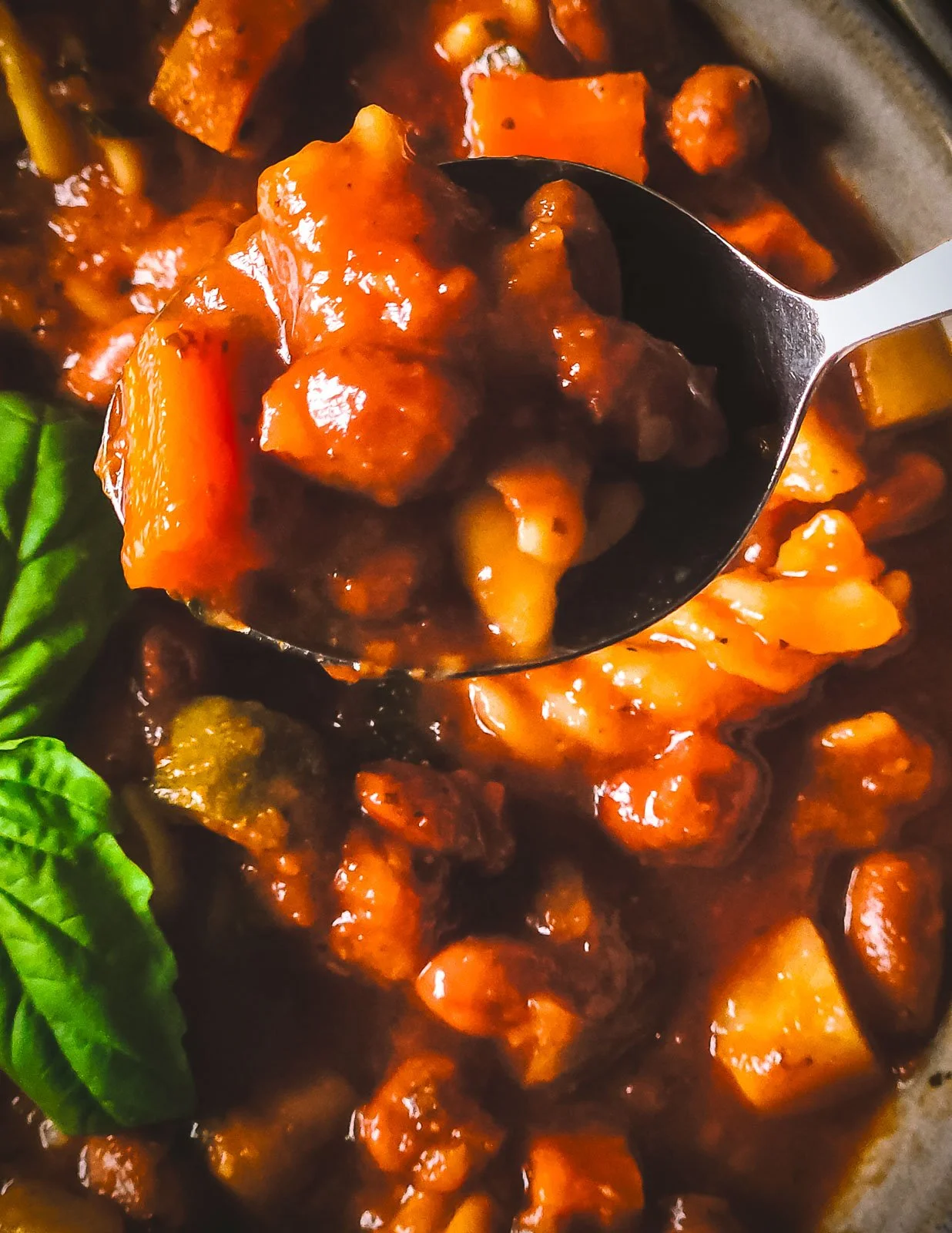 Close-up of a spoon hovering over a bowl of vegetarian minestrone soup, showing carrots, potatoes, beans, courgette, and basil.