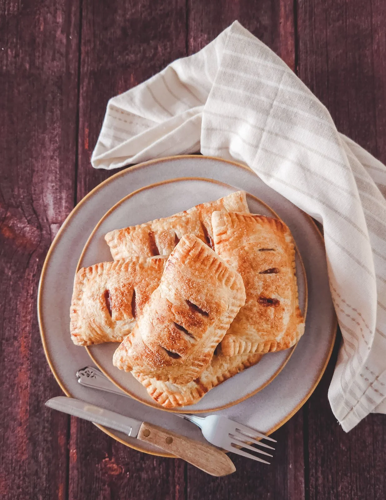 Rectangular apple puff pastry hand pies stacked on small plates over a larger plate, with a napkin and knife and fork on a wooden backdrop.