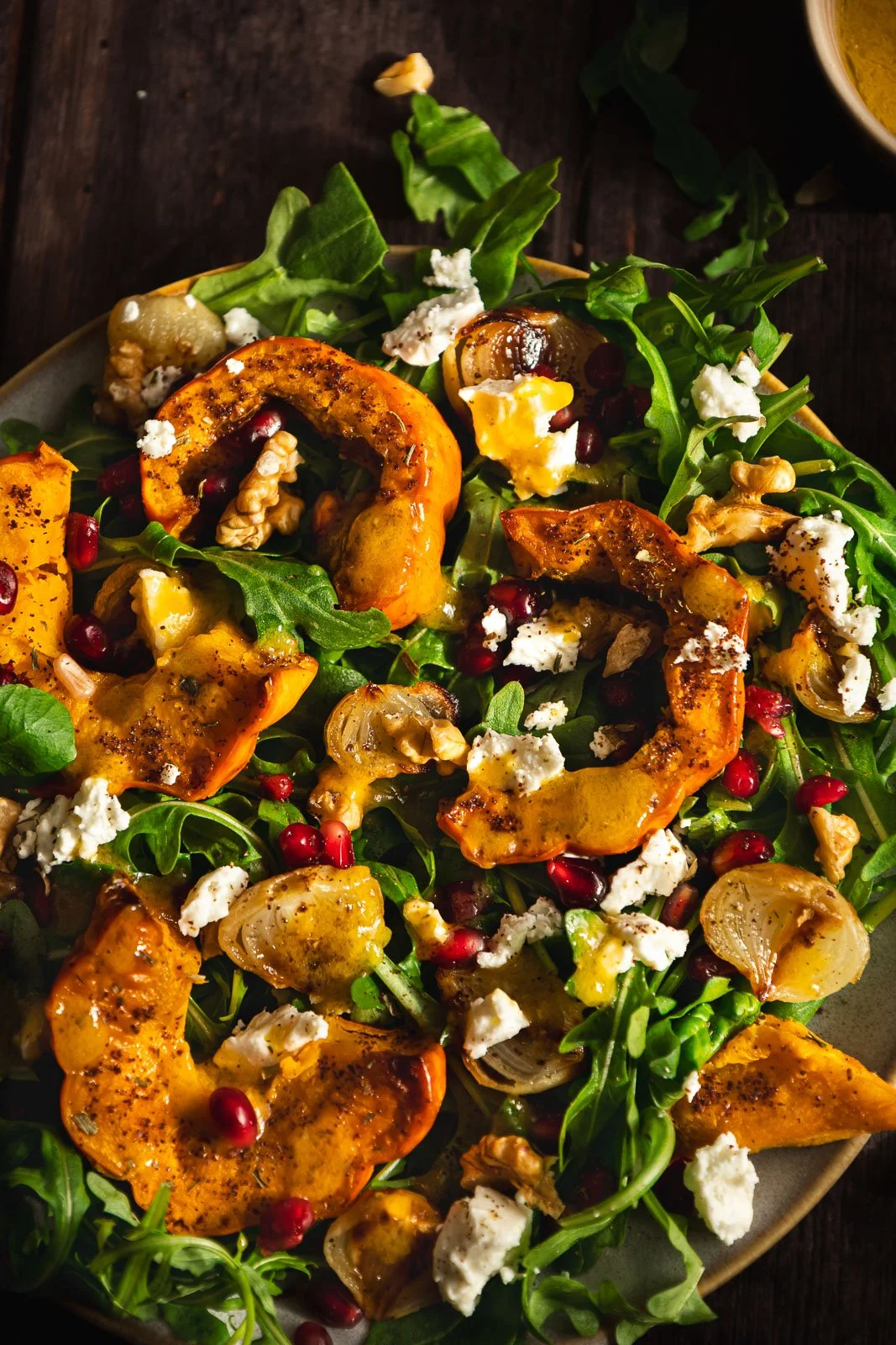 Close-up of roasted pumpkin with black pepper and herbs, crumbled feta, walnut pieces and glossy pomegranate seeds nestled among fresh rocket leaves.