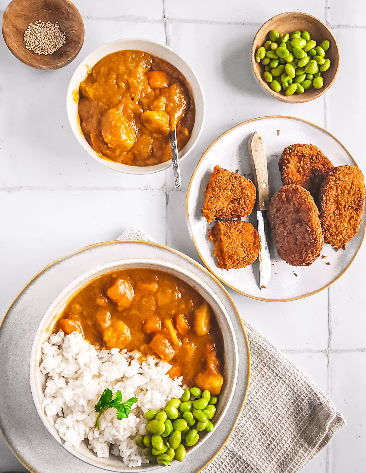 Flat lay of vegetarian Japanese curry with rice and edamame, served alongside pumpkin croquettes, sesame seeds, and a wooden bowl of edamame on a tiled background.