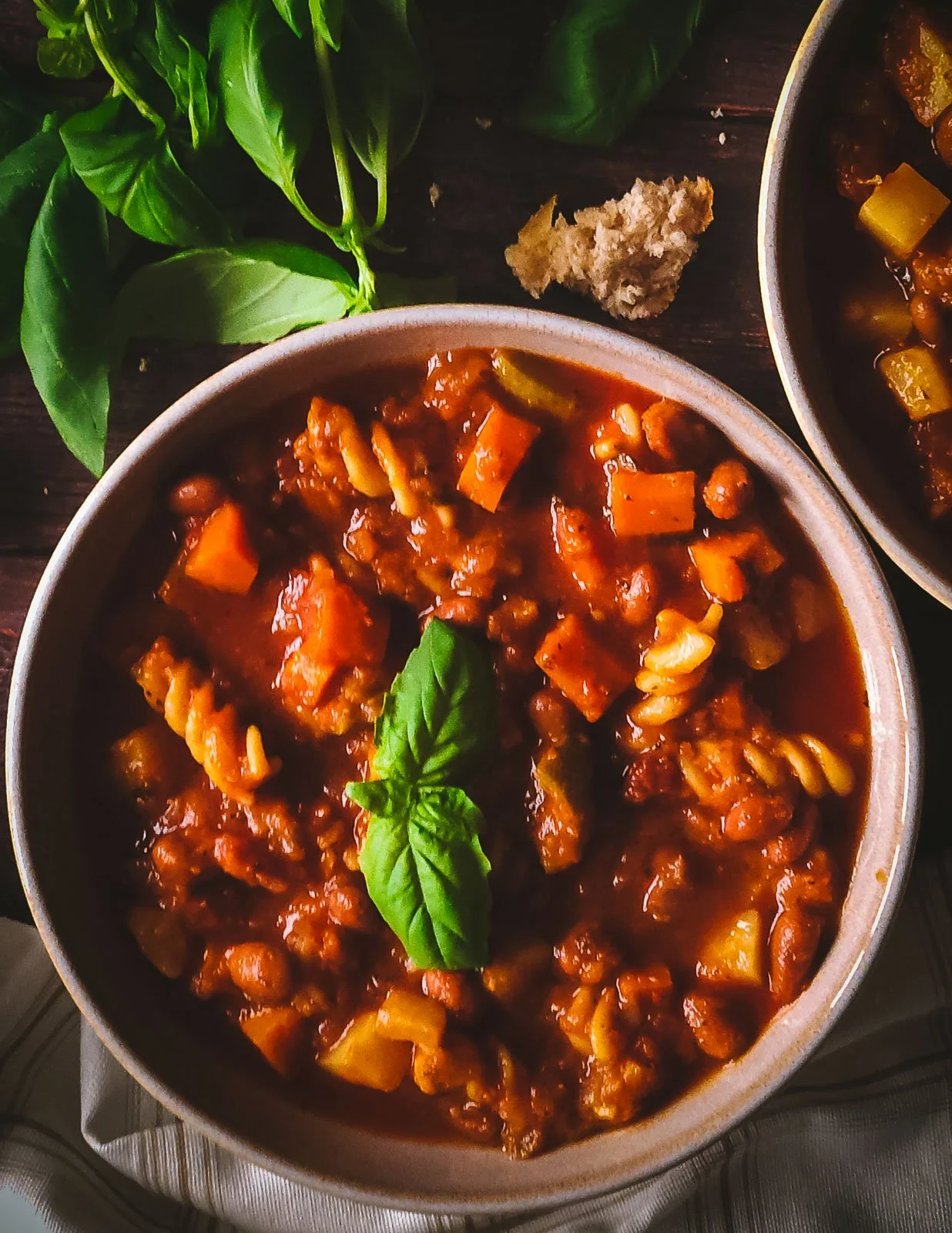 Close-up flat lay of minestrone soup with basil garnish and rustic bread pieces, styled with a napkin on a dark wooden background.
