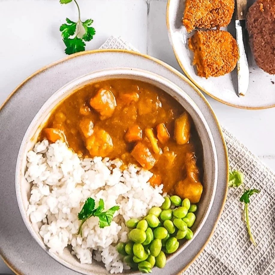 Beige bowl of vegetarian Japanese curry with rice and edamame beans, garnished with a single coriander leaf, set on a beige plate with a napkin on a white tiled background.