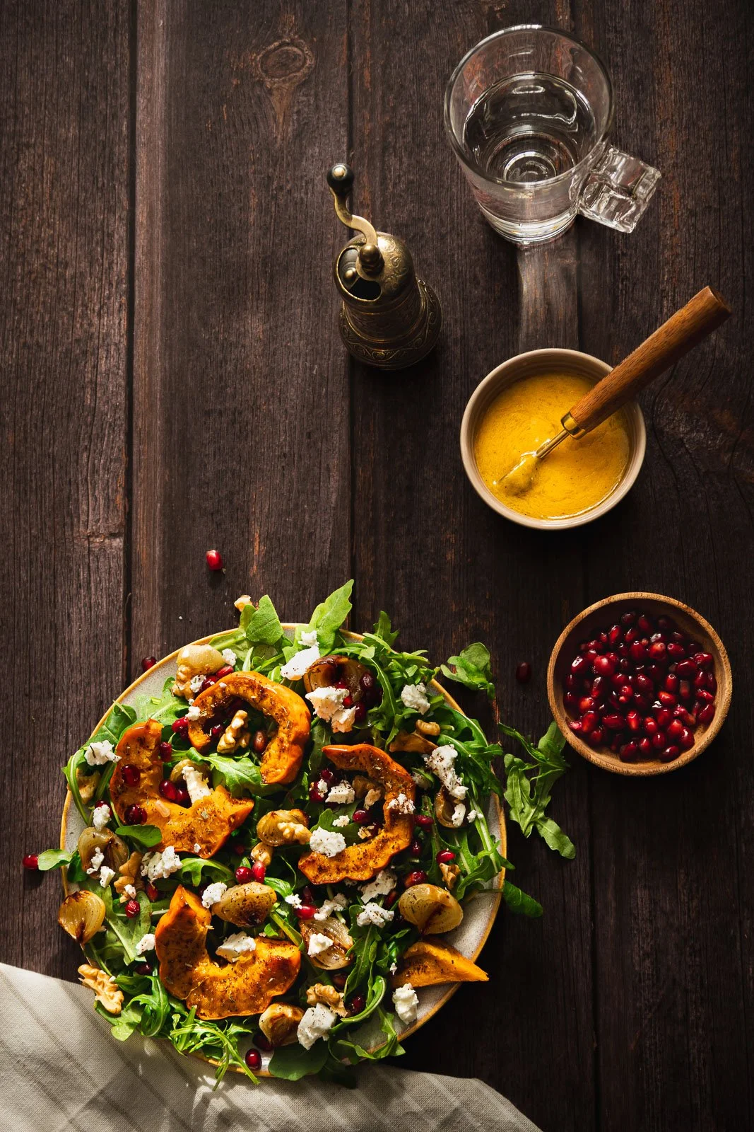 Overhead view of roasted munchkin pumpkin and baby onion salad with rocket, crumbled feta, walnuts and pomegranate, served on a ceramic plate with peach vinaigrette and a glass of water.