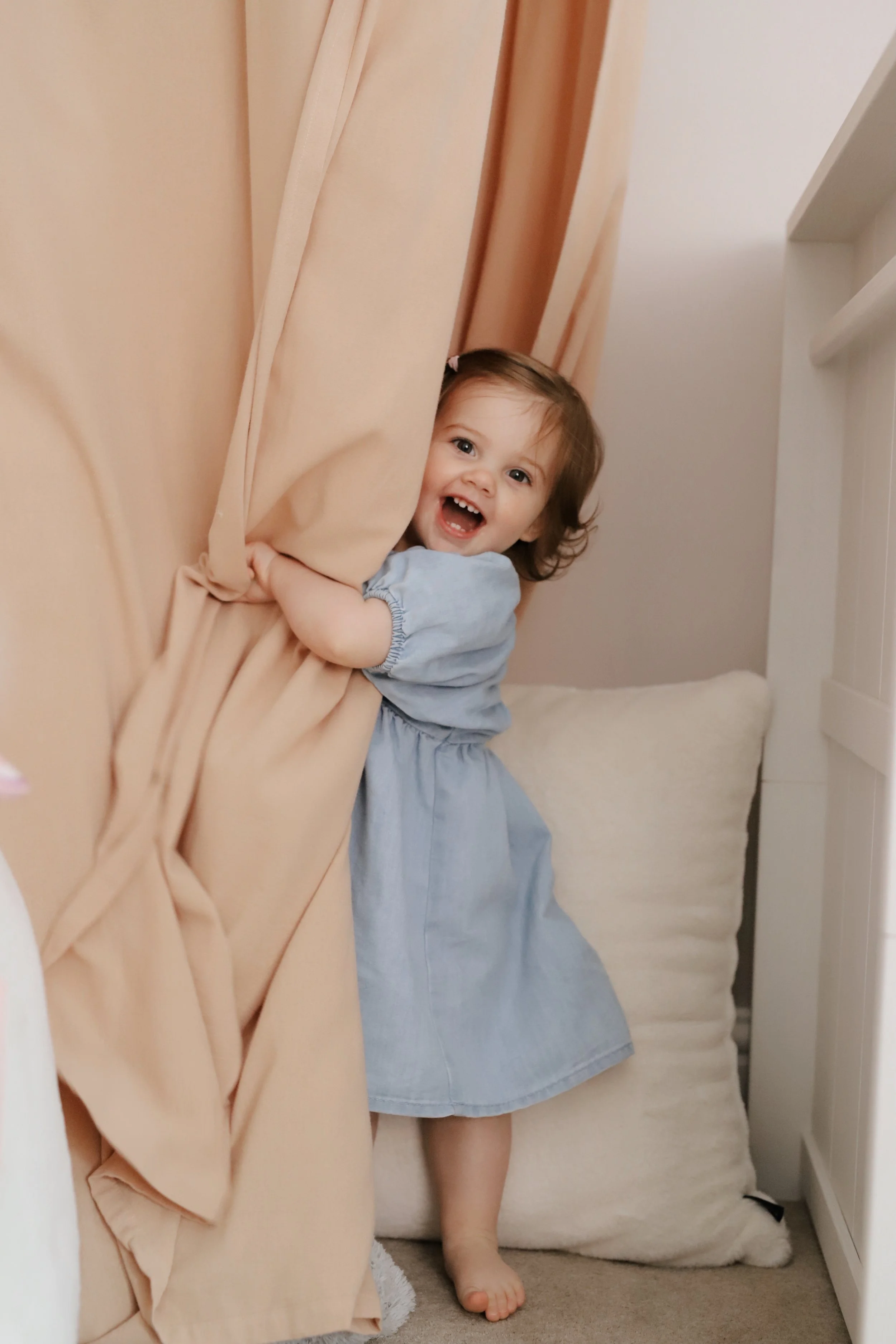 A young girl with brown hair and a big smile, wearing a light blue dress, peeking out from behind a beige curtain in a cozy indoor space.