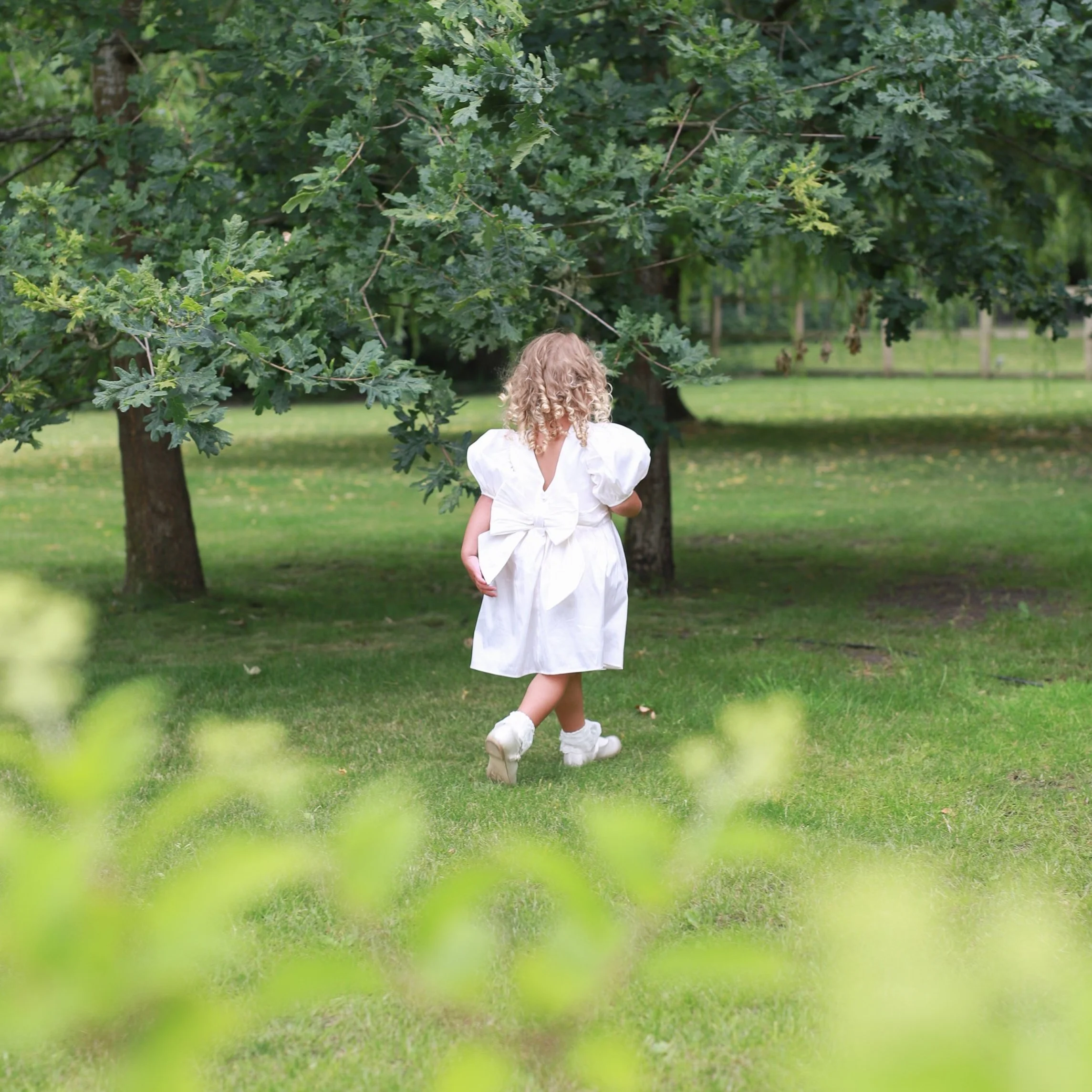 A young girl with curly blonde hair wearing a white dress with puffed sleeves and a big bow back, walking in a park with green grass and trees.