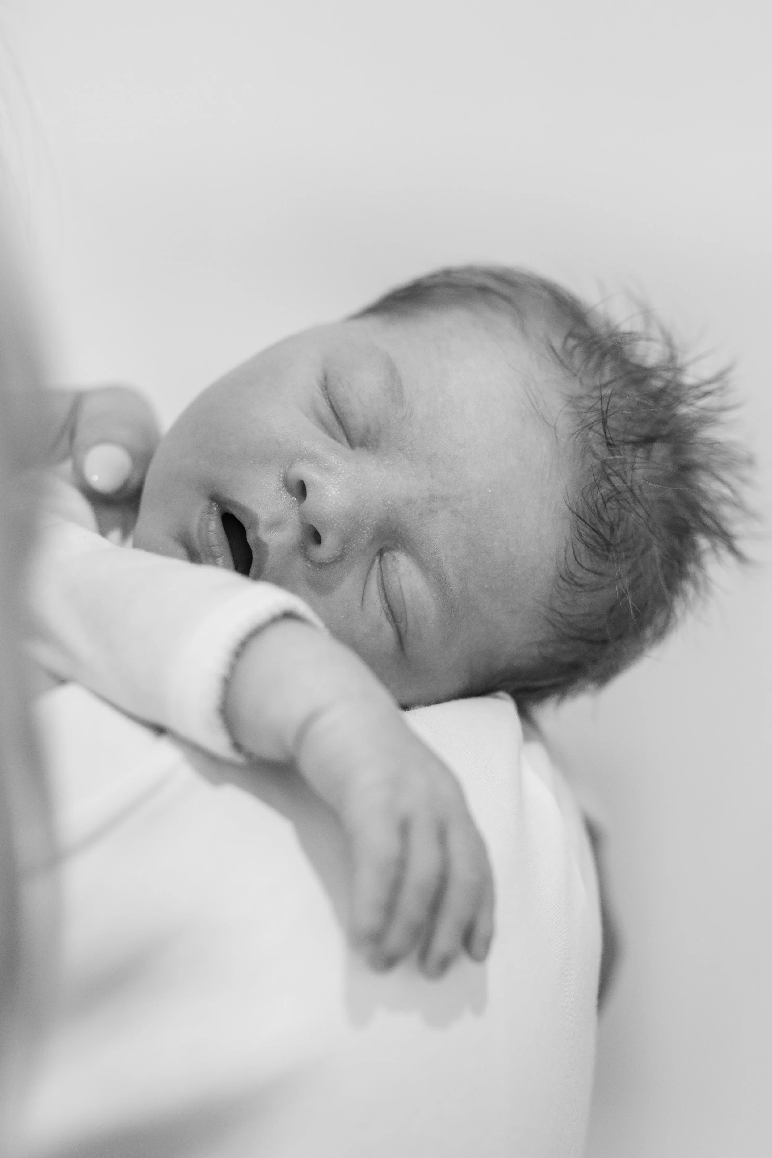 Black and white photo of a newborn baby sleeping on white blankets, with just a hint of a caregiver's hand visible in the frame.