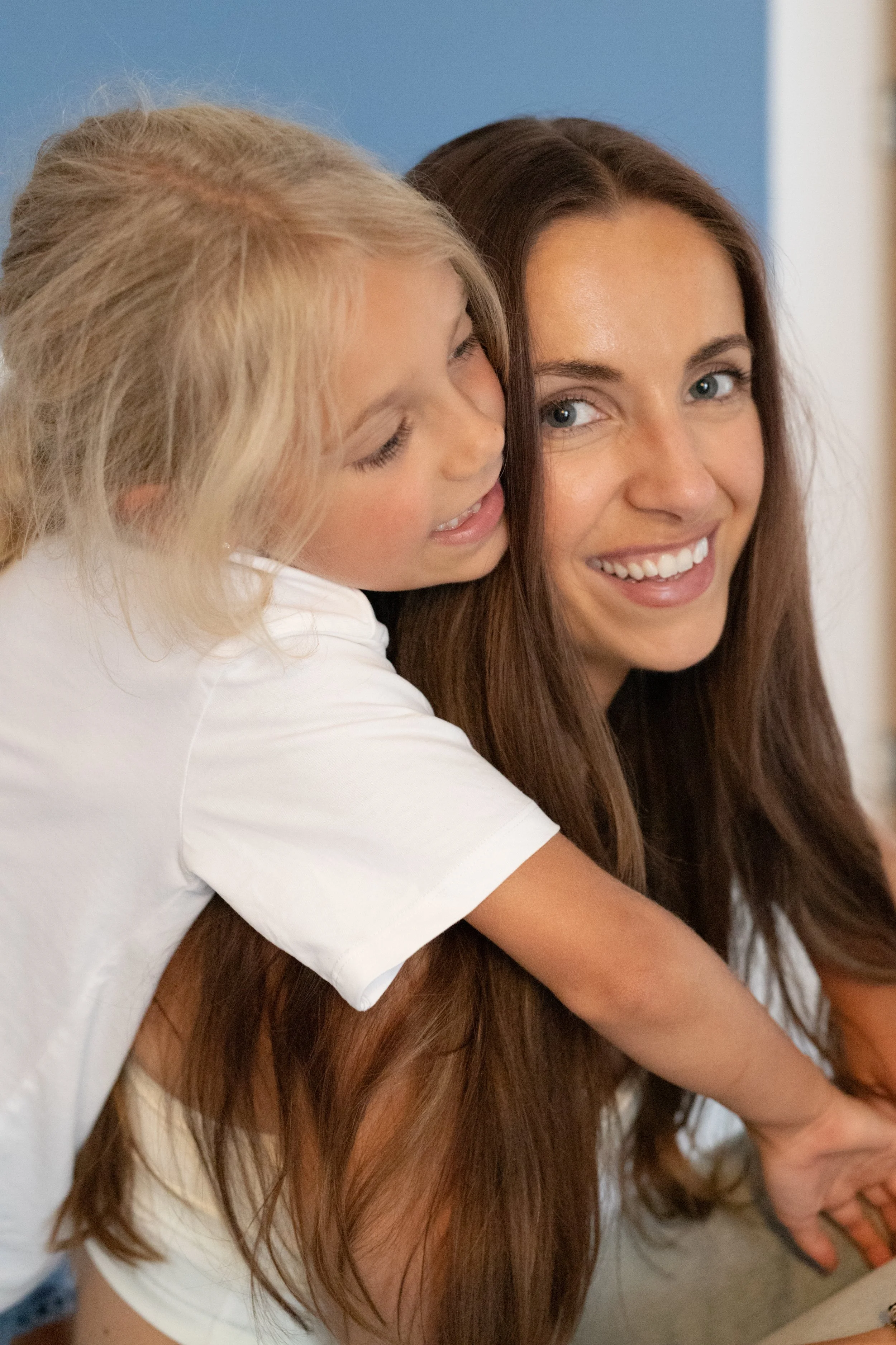 A woman with long brown hair and blue eyes smiling while a young girl with blonde hair and closed eyes hugs her from behind.